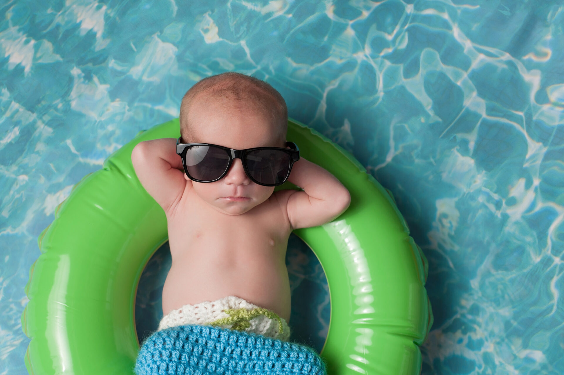 Infant floating on green pool ring wearing oversized sunglasses in clear blue water