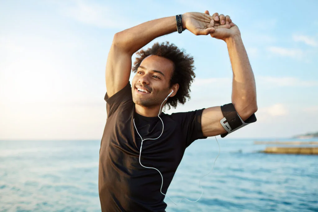 Man stretching by the ocean wearing black shirt and earbuds with armband