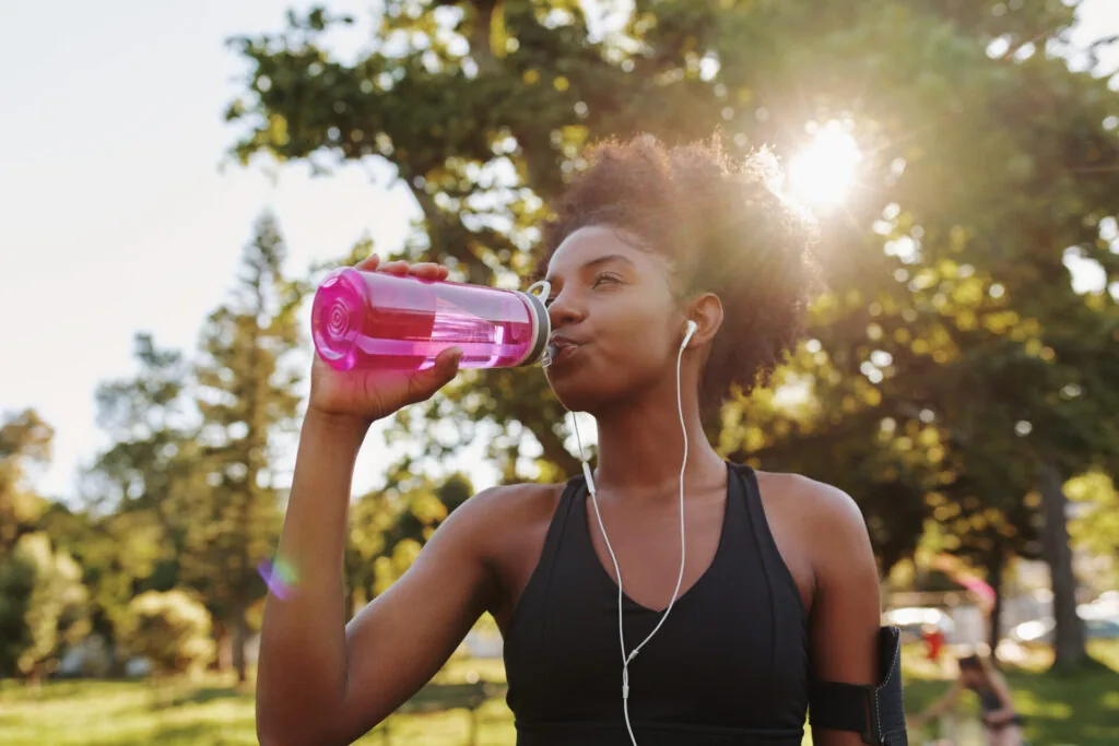 Woman in black sports top drinking from pink water bottle while wearing earbuds in sunny park