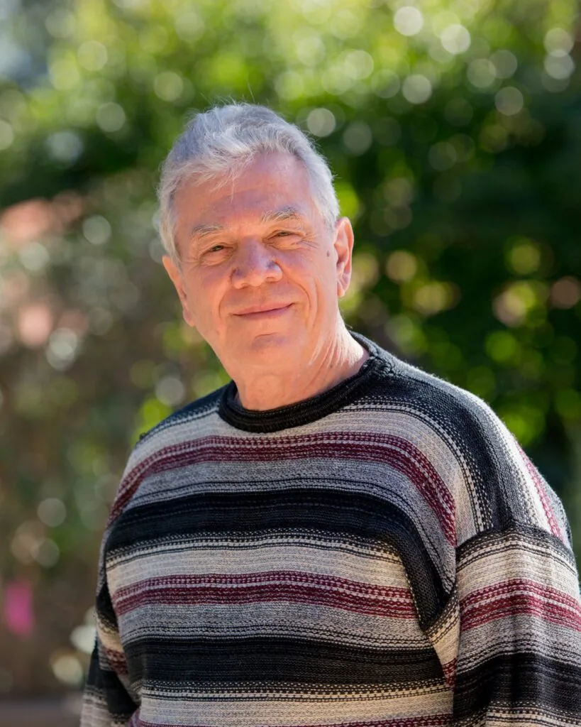 Older man with white hair wearing a striped sweater outdoors with blurred green background