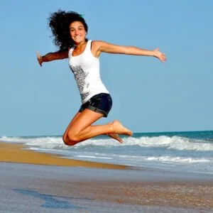 Woman jumping joyfully on beach with arms outstretched, ocean and waves in background under clear blue sky