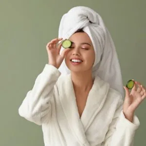 Woman in white bathrobe and towel-wrapped hair holding cucumber slices over eyes against green background